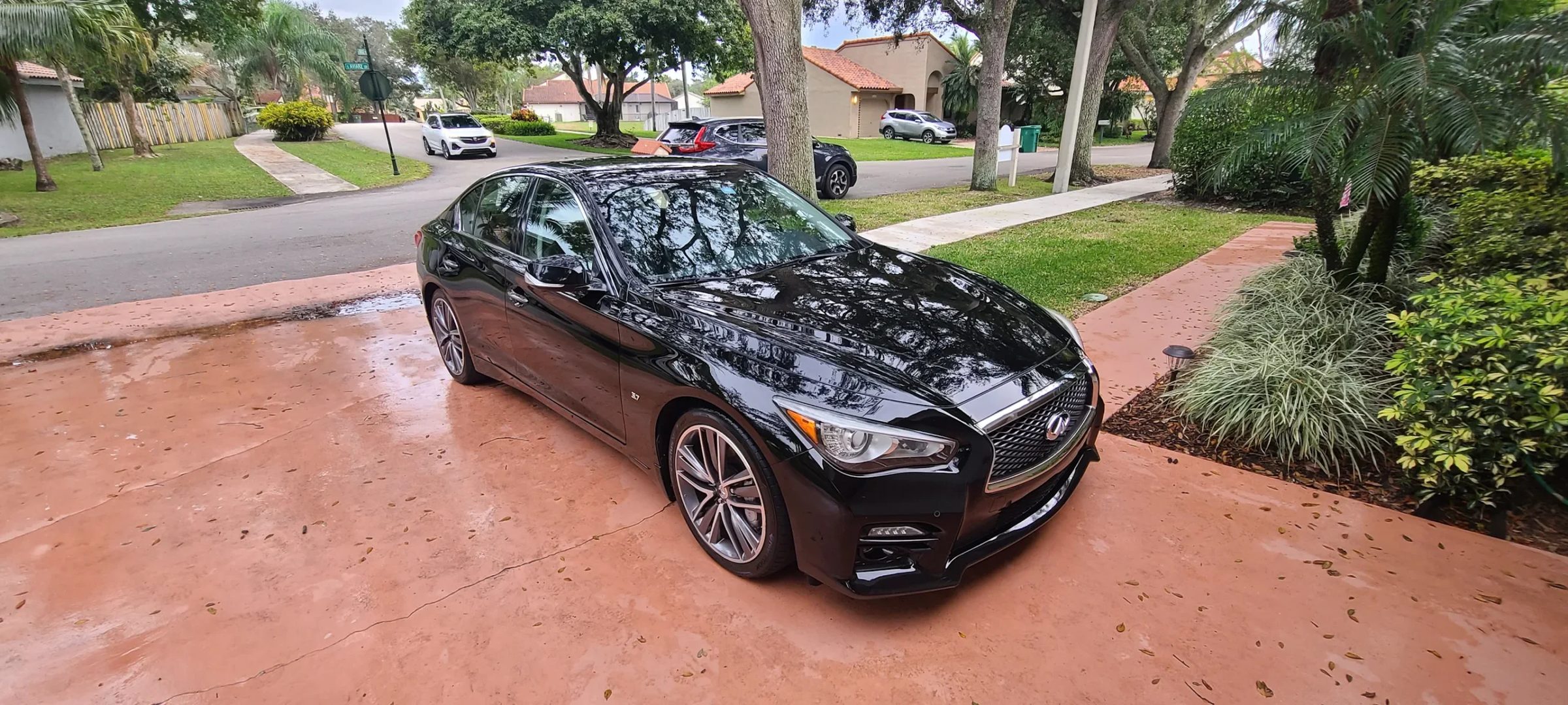 Dark Infiniti Q50 sedan after a full hand wash, parked on a wet red paver driveway in a palm-lined neighbourhood