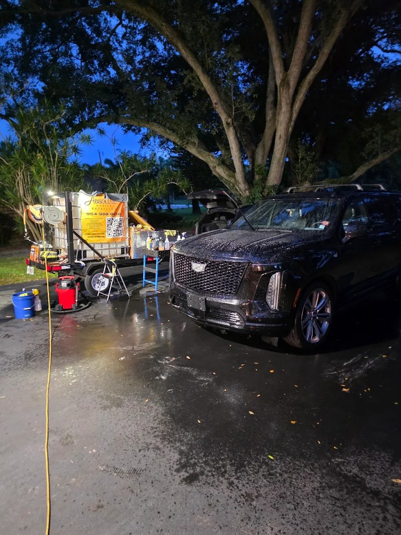 Black Cadillac Escalade being washed after dark under work lights, hoses and buckets laid out beside the detailing trailer